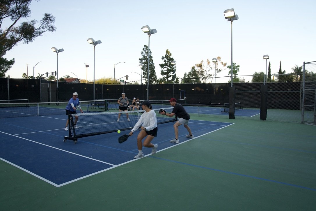 People playing pickleball on an outdoor court with blue and green surfaces, surrounded by a black fence and tall lights, during sunset.