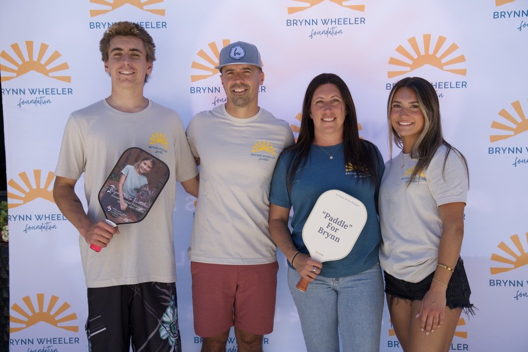Four people standing in front of a Brynn Wheeler Foundation backdrop, smiling, holding signs, with three wearing relative t-shirts, one in a teal t-shirt, and one in a white t-shirt.