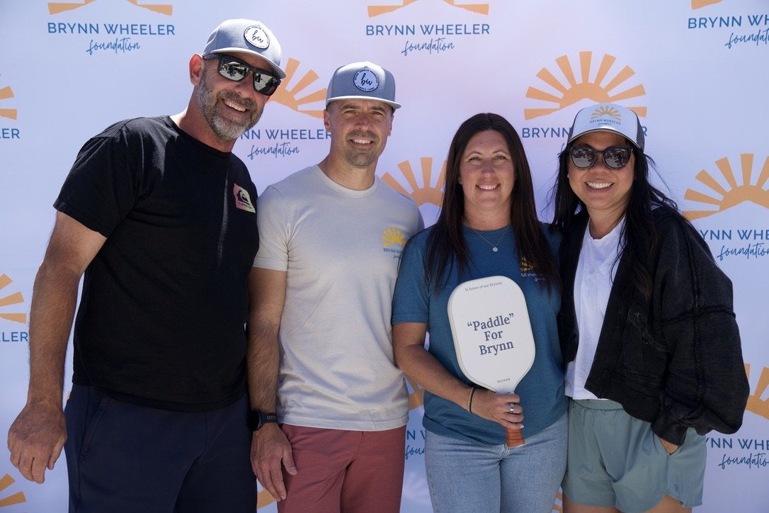 Four people standing outdoors at a Brynn Wheeler Foundation event, smiling, with a backdrop featuring the foundation's logo. One woman holds a paddle labeled 'Paddle for Brynn.'