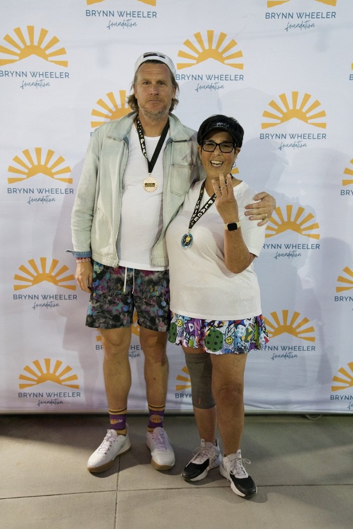 A man and a woman standing together in front of a Brynn Wheeler Foundation backdrop, both wearing medals. The man is dressed in a white jacket, shorts with a colorful pattern, and sneakers, with a cap and several bracelets. The woman is wearing a whi