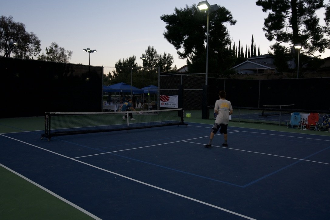 Two people playing pickleball on an outdoor court during early evening, with trees and a few tents in the background.