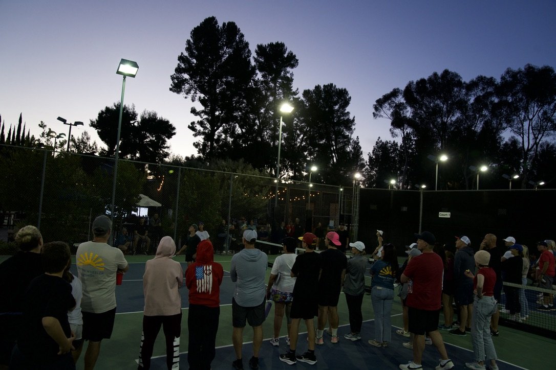 A group of people gathering on a tennis or pickleball court at dusk, with trees and bright overhead lights in the background.