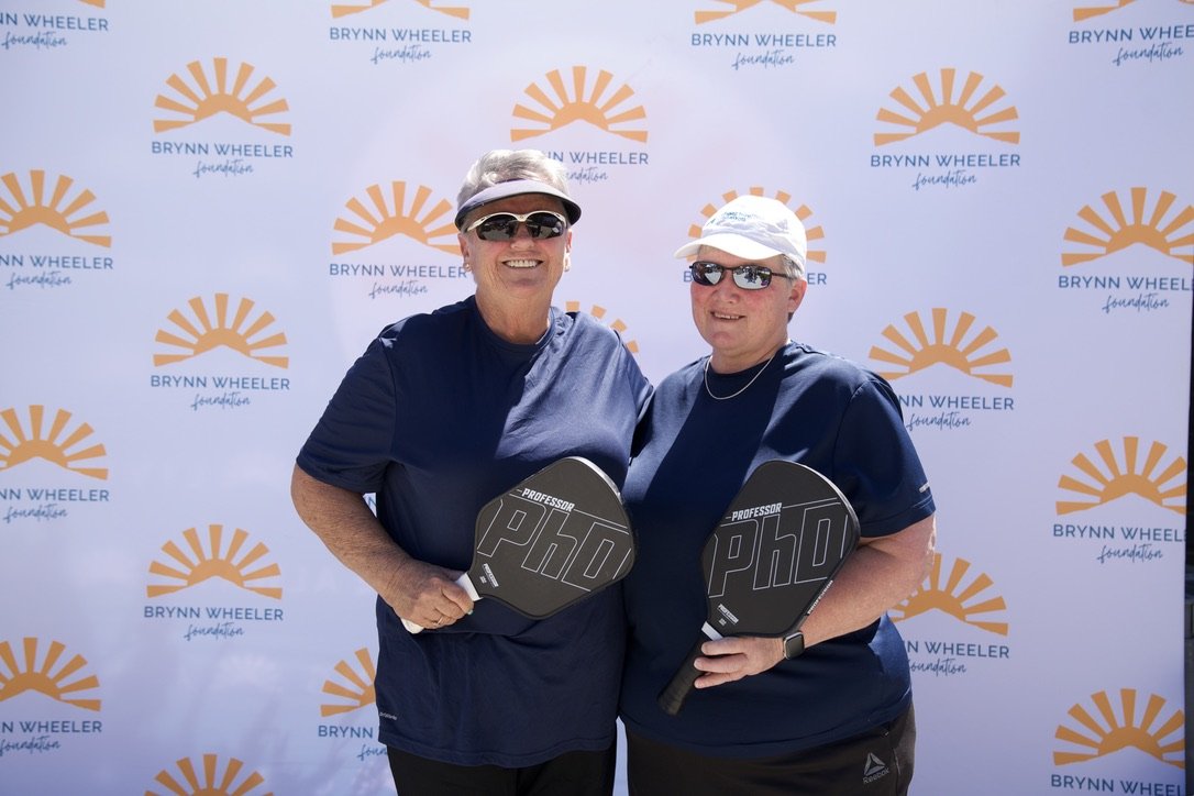 Two women standing together outdoors, holding pickleball paddles, smiling, in front of a backdrop with Brynn Wheeler Foundation logos.