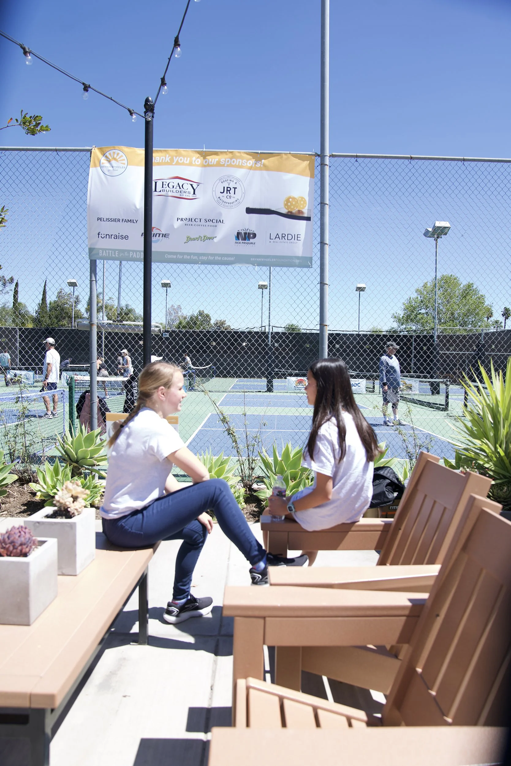 Two women sitting on benches near a tennis court, talking. The tennis court is behind a chain-link fence with a banner hanging, and people are playing tennis on a sunny day.