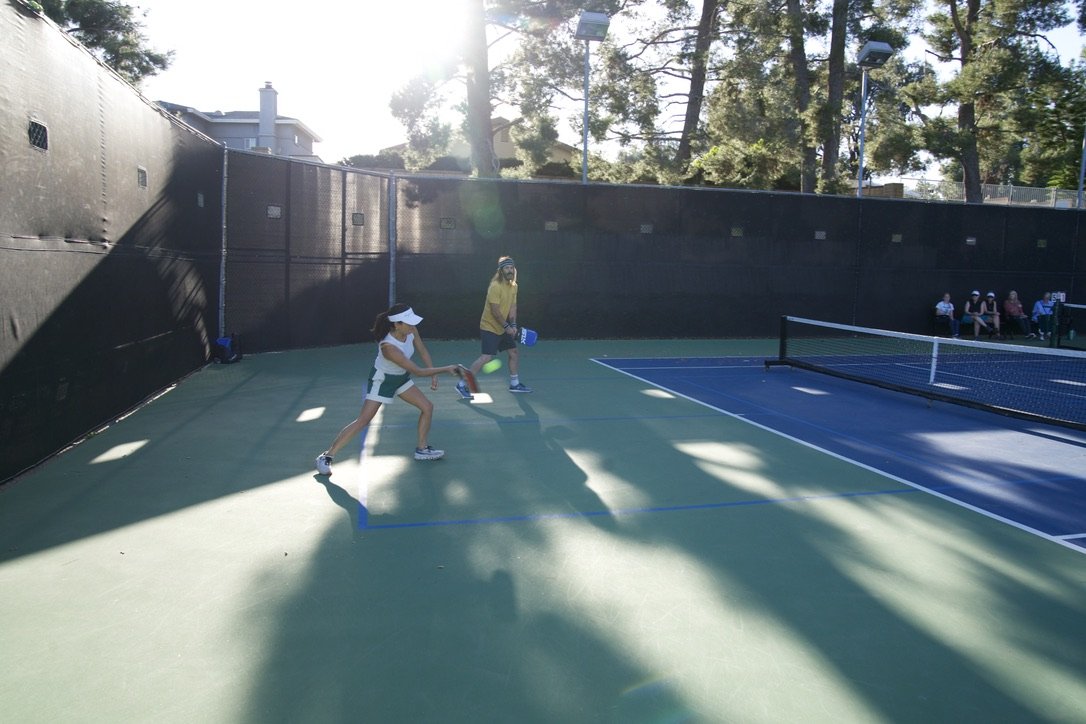 Two young girls playing pickleball on an outdoor court with an adult nearby. There are spectators sitting on benches in the background and trees surrounding the court.
