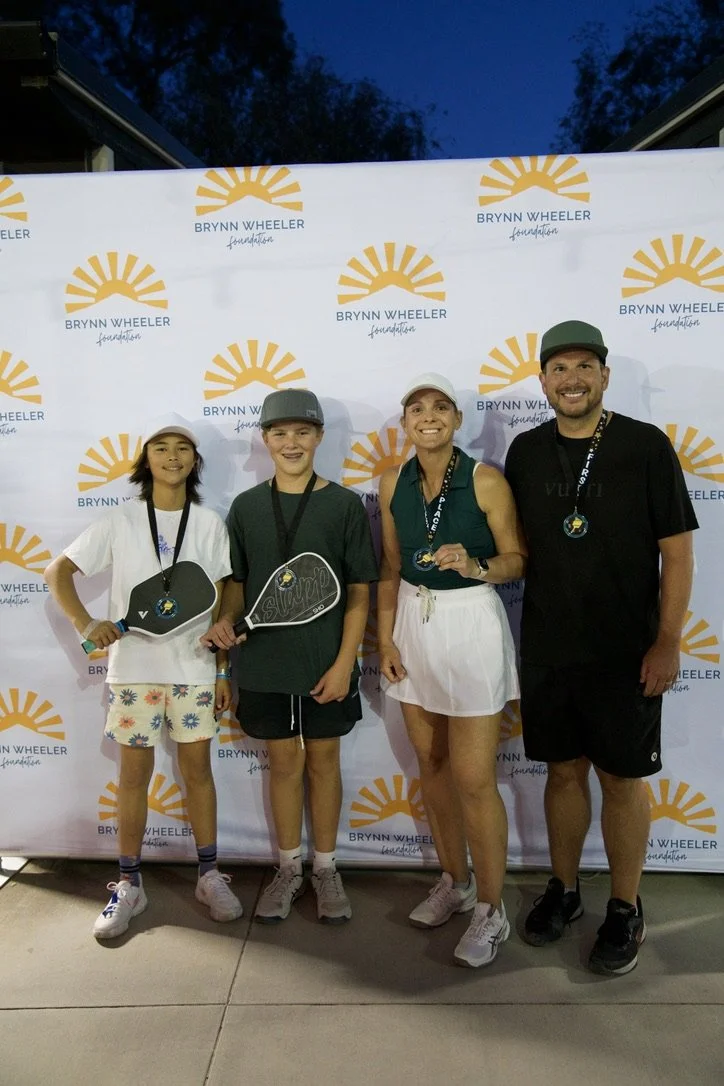 Four people standing in front of a Brynn Wheeler Foundation backdrop, holding medals and sports props, smiling.
