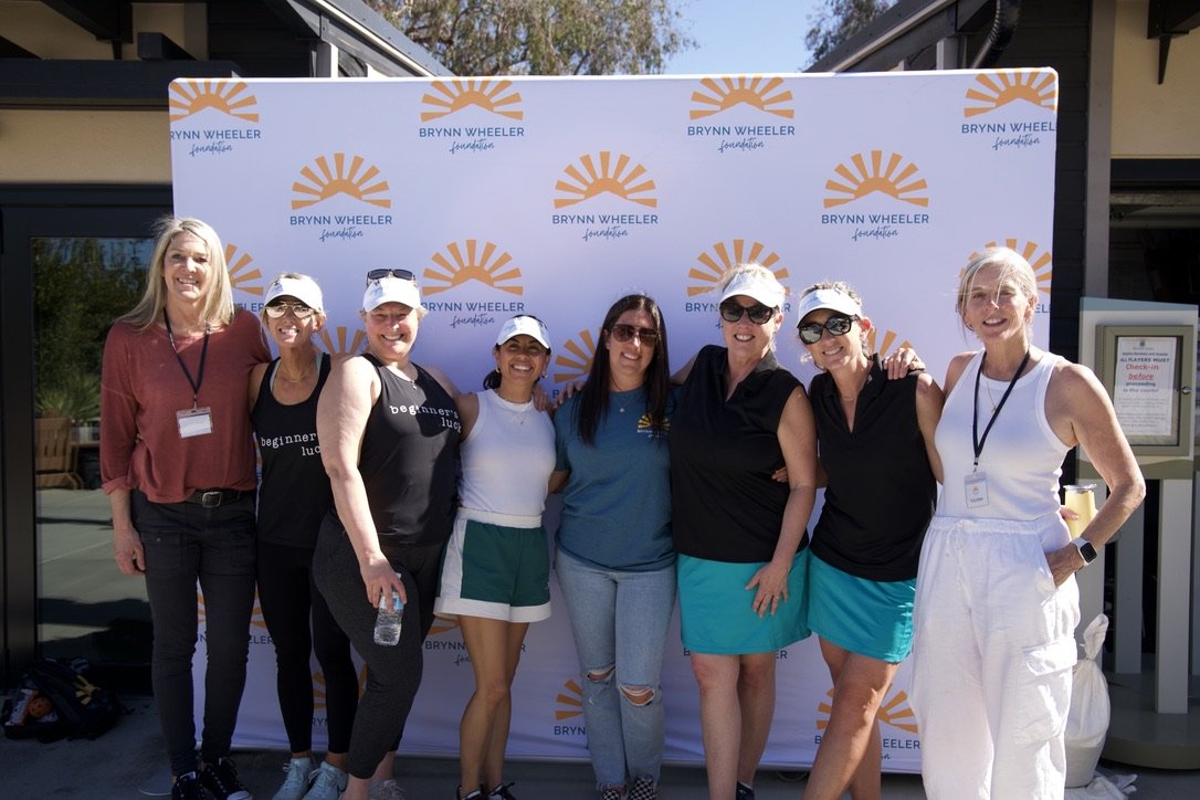 Group of women standing in front of a Brynn Wheeler Foundation backdrop, smiling, outdoors on a sunny day.