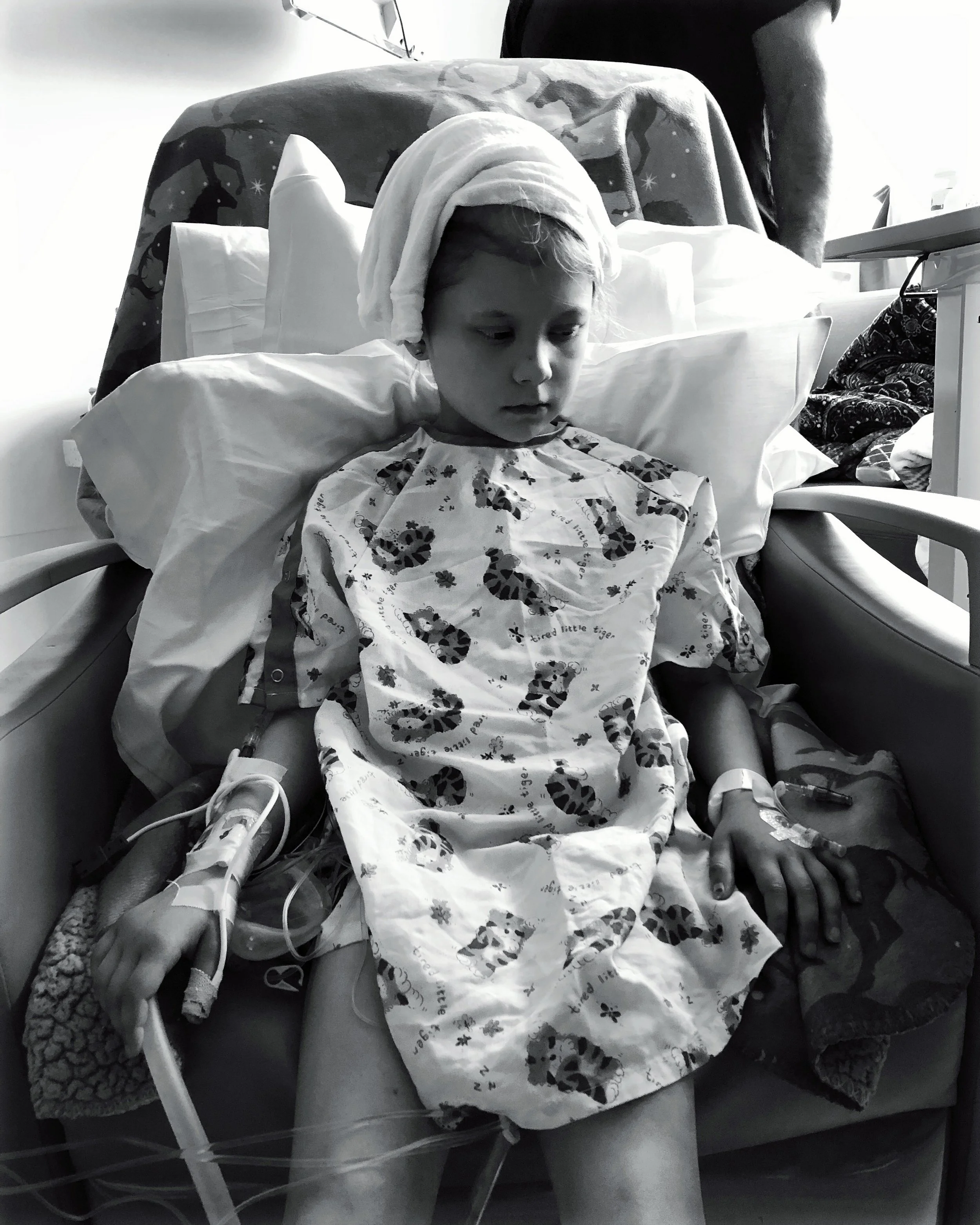 Child sitting in a hospital chair, wearing a patterned gown and head wrap, with an IV in their arm and a hospital bracelet visible. Black and white photograph.