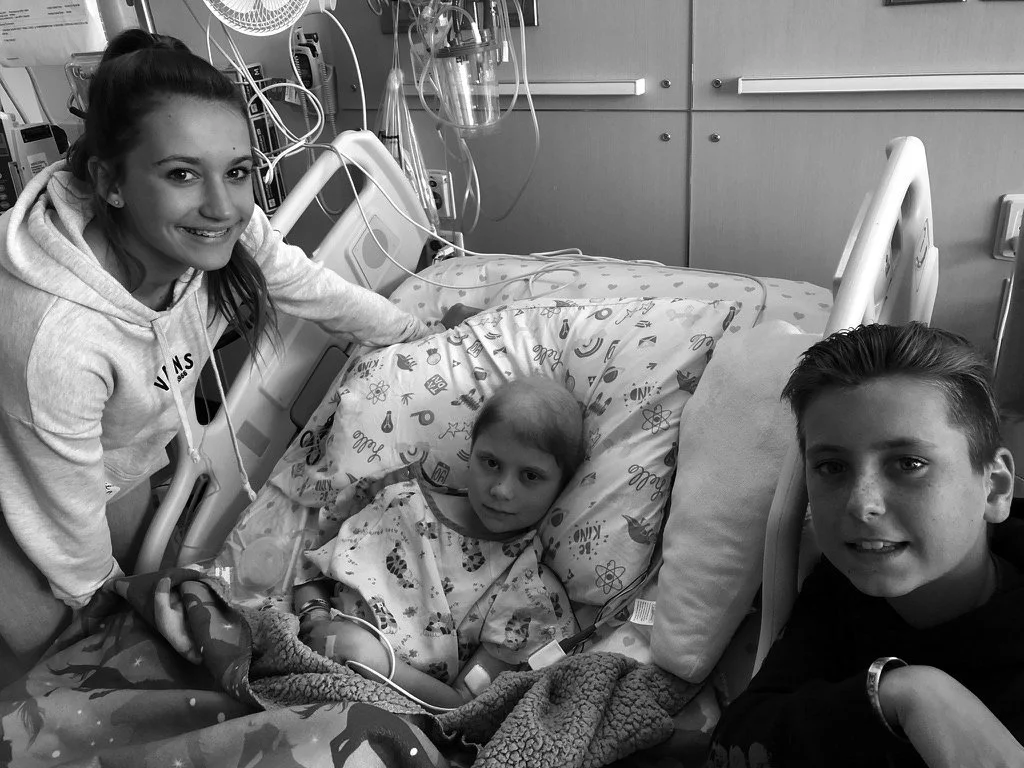 Three children in a hospital setting, with one child lying in a hospital bed connected to medical equipment, accompanied by two other children beside the bed.