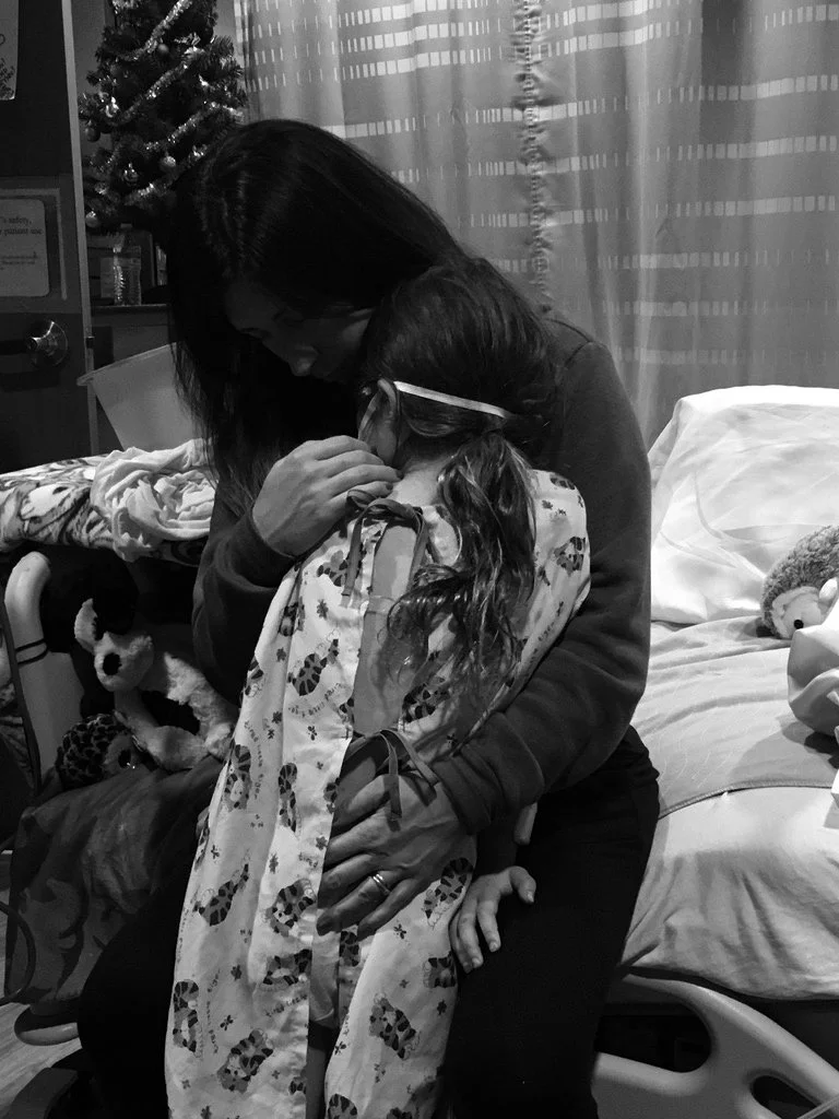 A black and white photo showing a woman holding a young girl wearing a hospital gown, sitting on a bed in a room with a Christmas tree in the background.