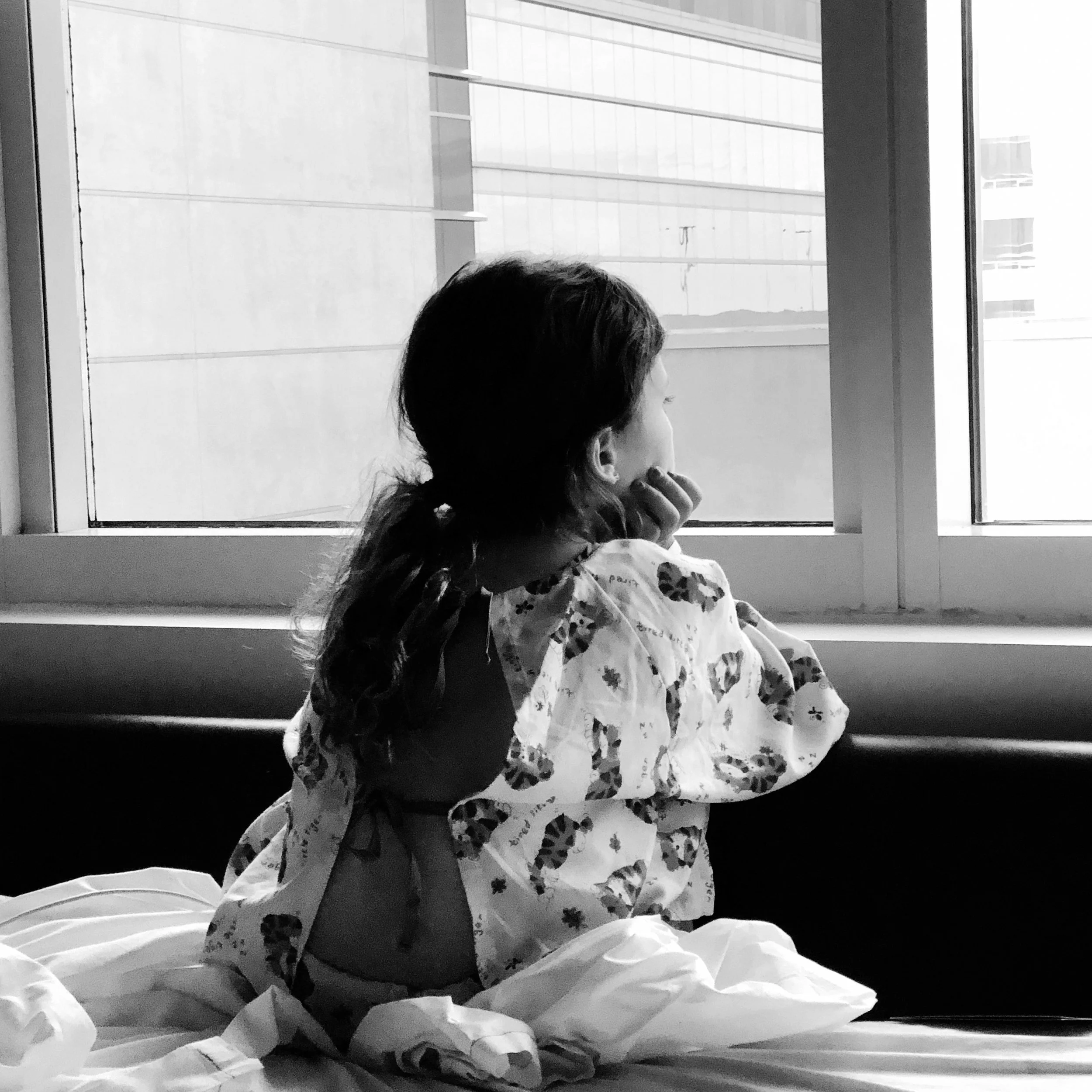 Black and white photo of a child wearing a hospital gown, looking out of a window, seated on a bed.