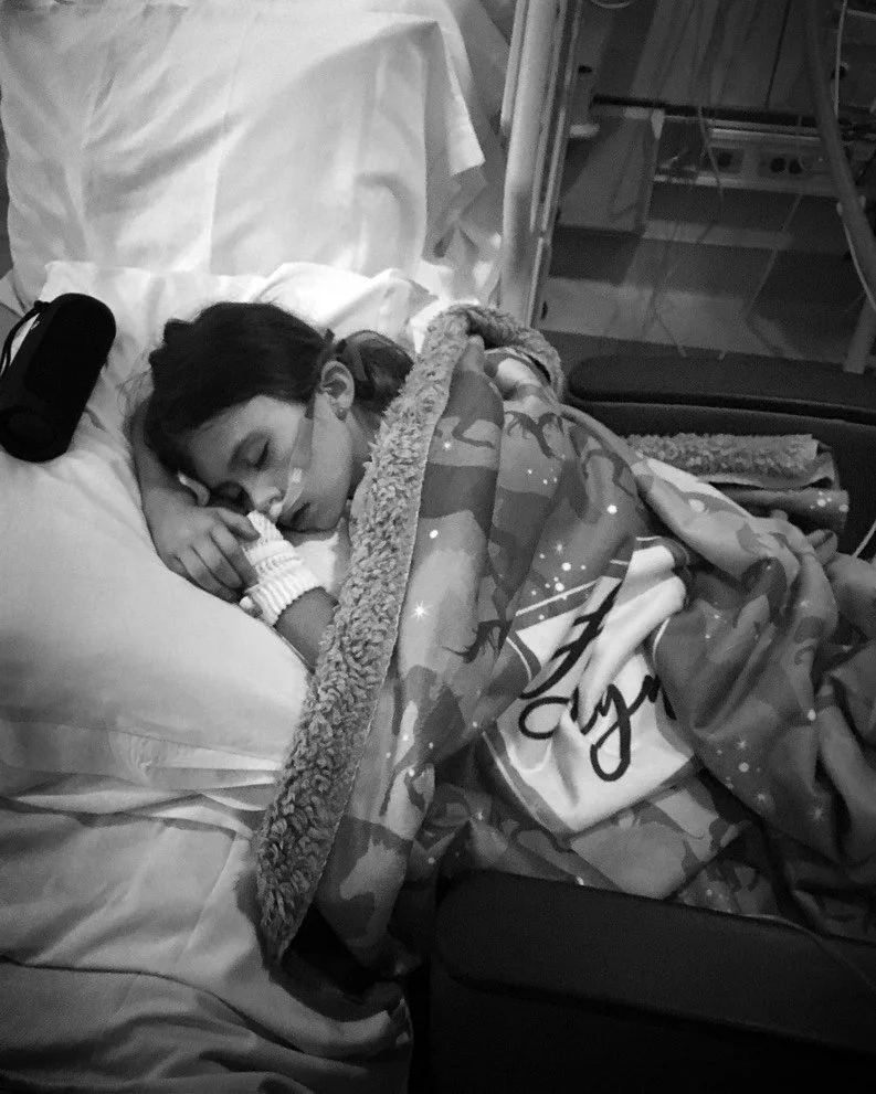 Child sleeping in a hospital bed, covered with a patterned blanket and wearing a nasal oxygen tube.