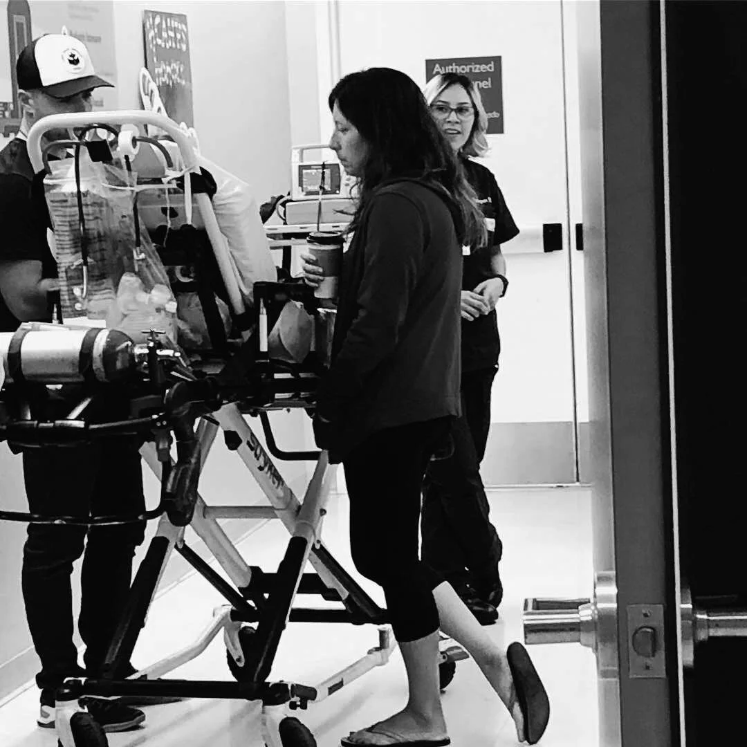 Black and white image of three people standing near a hospital stretcher in a hallway. One person is wearing a cap, another has a coffee cup, and a third is dressed in medical attire.