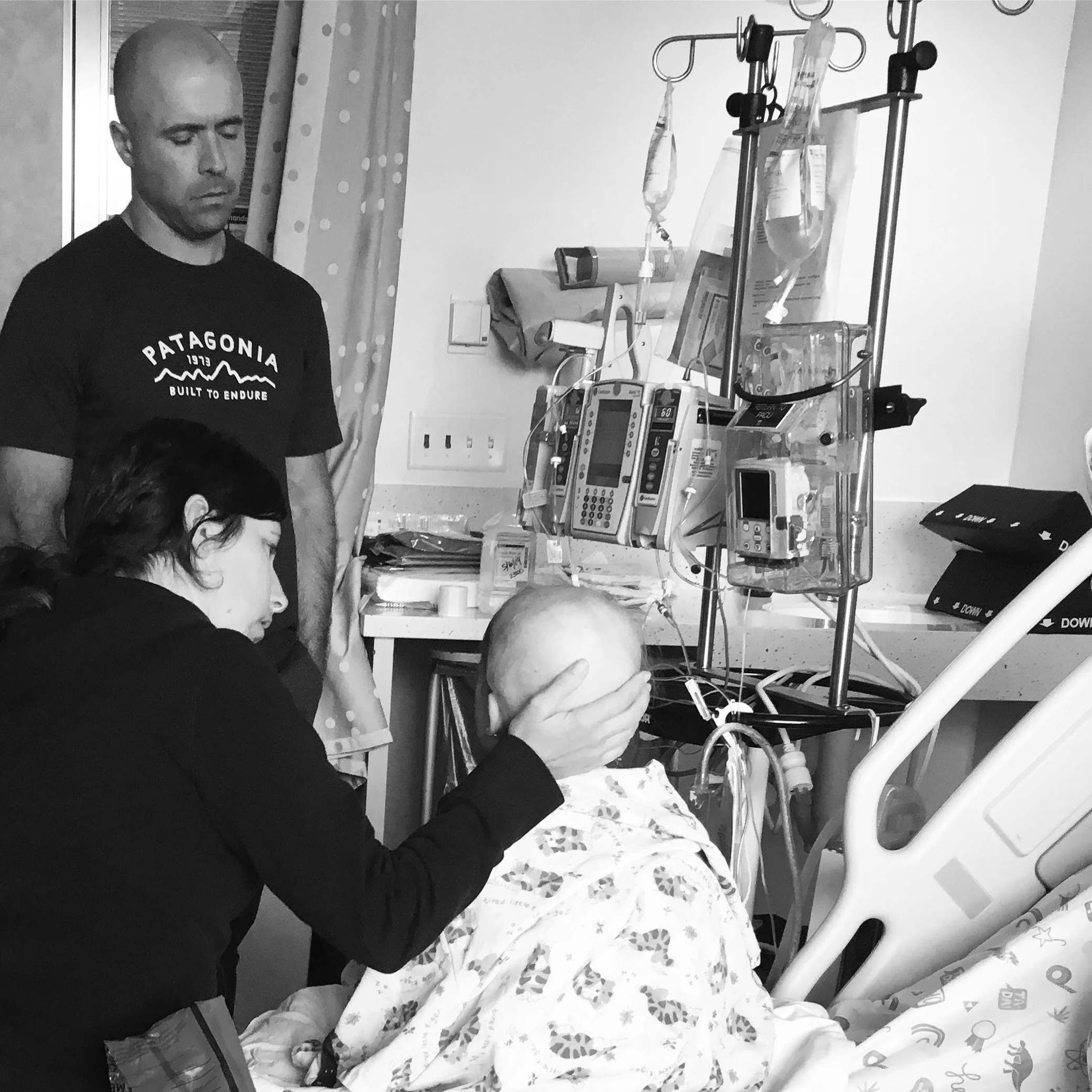 A hospital room scene showing a patient in a hospital gown sitting on a bed with IV drips and medical equipment nearby. A woman is gently supporting the patient's head, and a man stands nearby with a concerned expression.