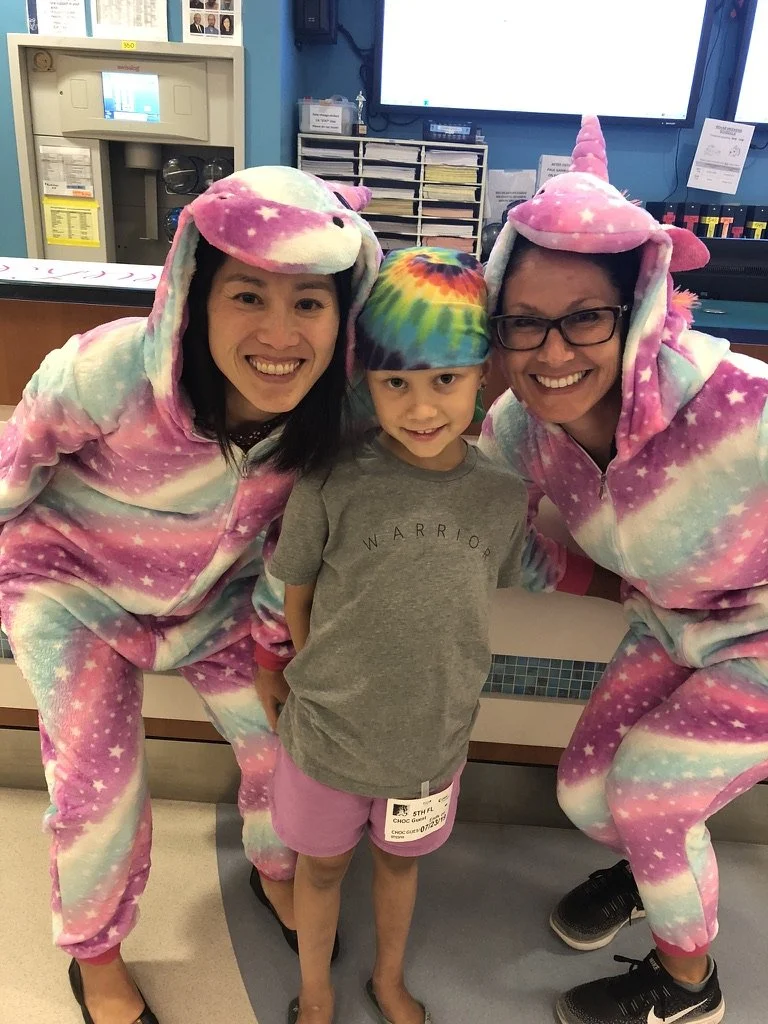 Two adults in unicorn onesies pose with a child wearing a tie-dye cap and gray shirt that says 'Warrior.'