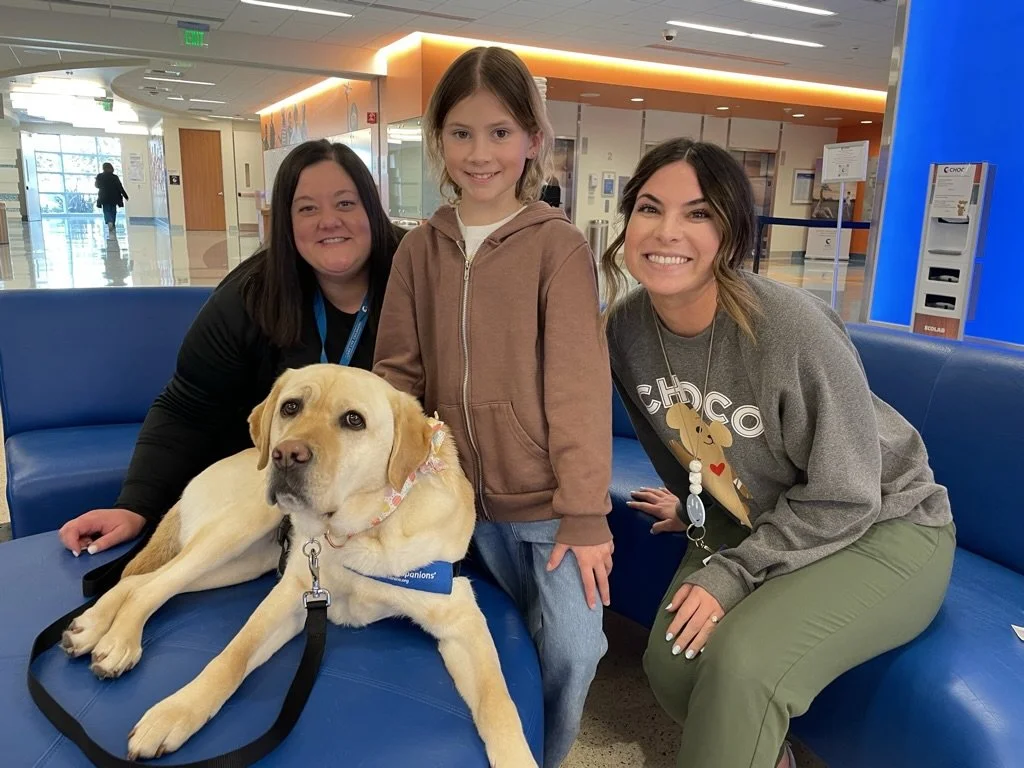 A group of three people sitting on a blue couch with a yellow Labrador retriever. The dog is wearing a collar and laying down. The people are smiling, indoors in a bright, modern space.
