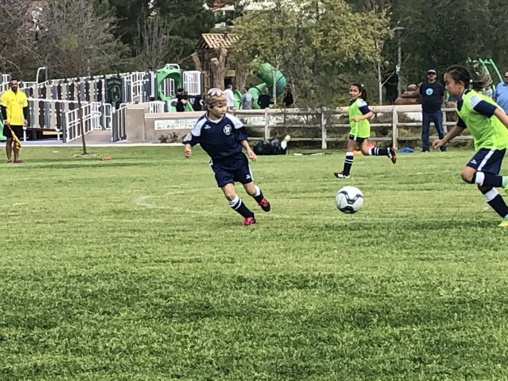 Youth soccer game with players in navy uniforms and green pinnies, playing on a grassy field with a playground in the background.
