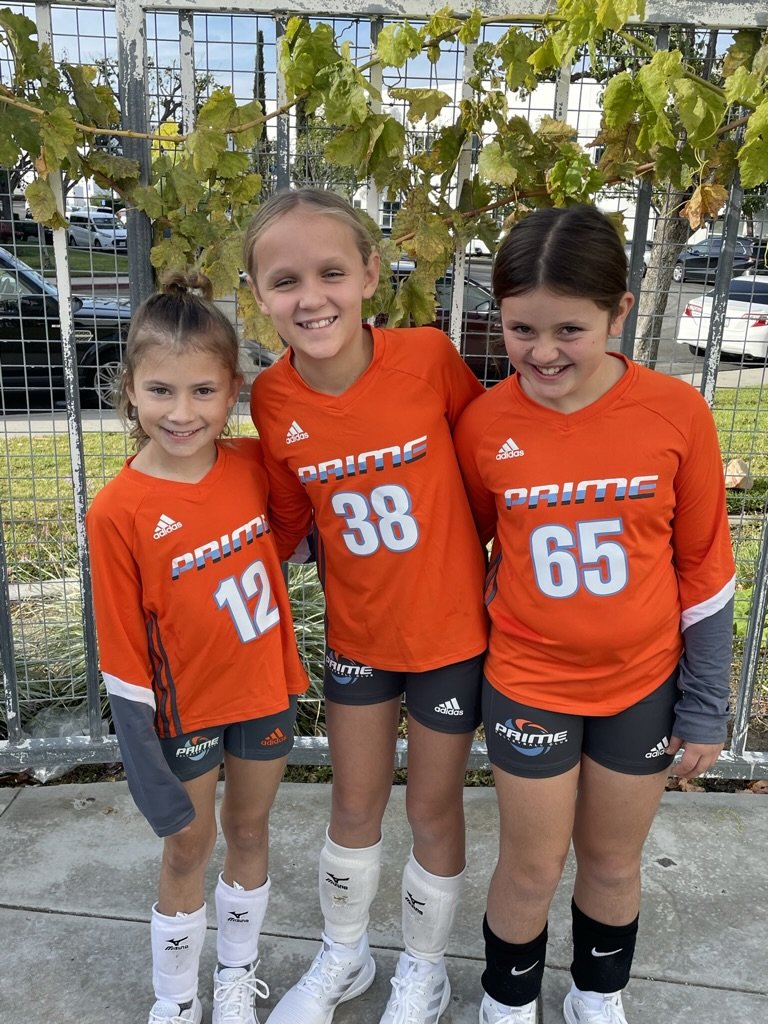 Three young girls in matching orange soccer uniforms, numbered 12, 38, and 65, standing together outside on a sidewalk with greenery in the background. They are wearing white knee socks and soccer shoes.