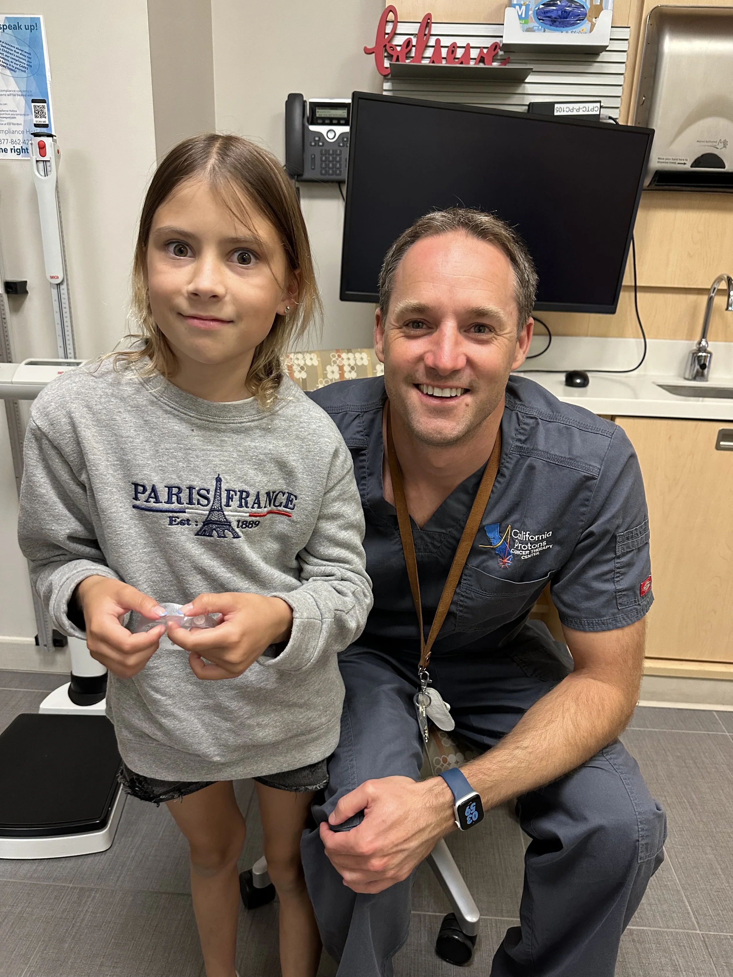 A young girl wearing a "Paris France" sweatshirt stands beside a man in medical scrubs, in what appears to be a doctor's office. The man is seated and smiling at the camera. In the background, there is a computer monitor and medical equipment.