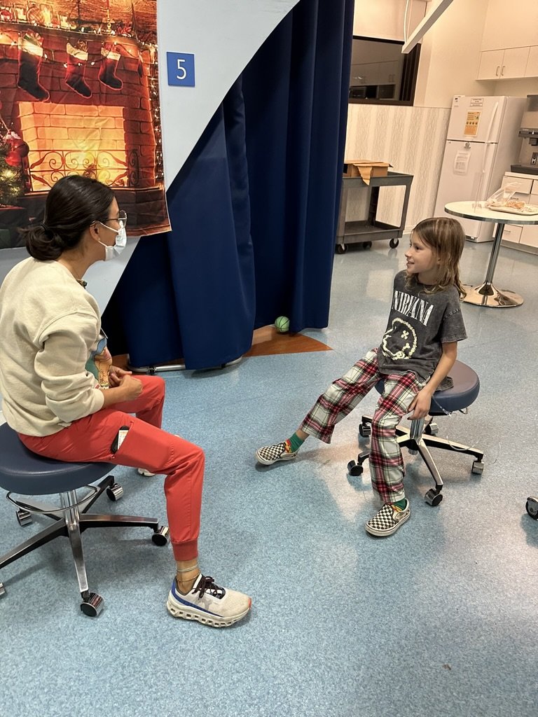 A person wearing red pants and a white sweater, sitting on a rolling chair while talking to a child in a dark T-shirt and plaid pants on another chair. There is a Christmas fireplace backdrop with stockings hanging, and a blue curtain with the number