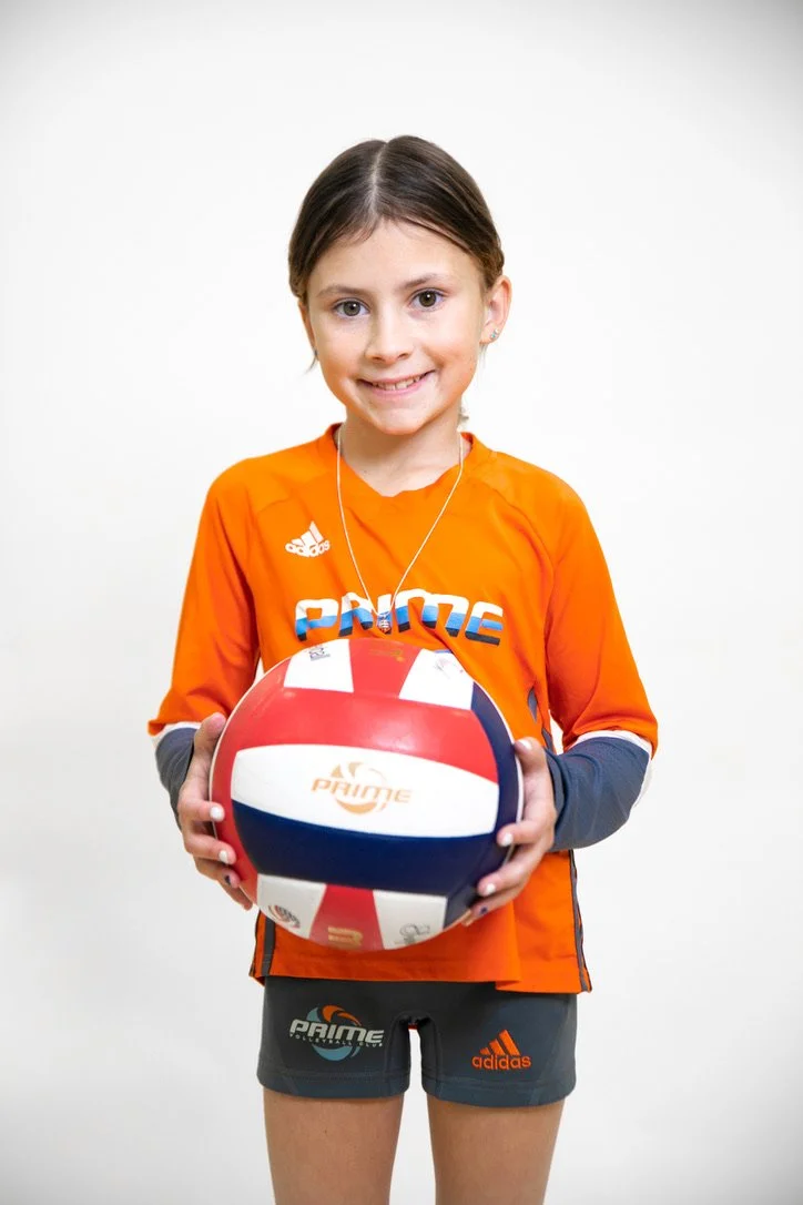 Young girl wearing an orange jersey and gray shorts holding a volleyball.