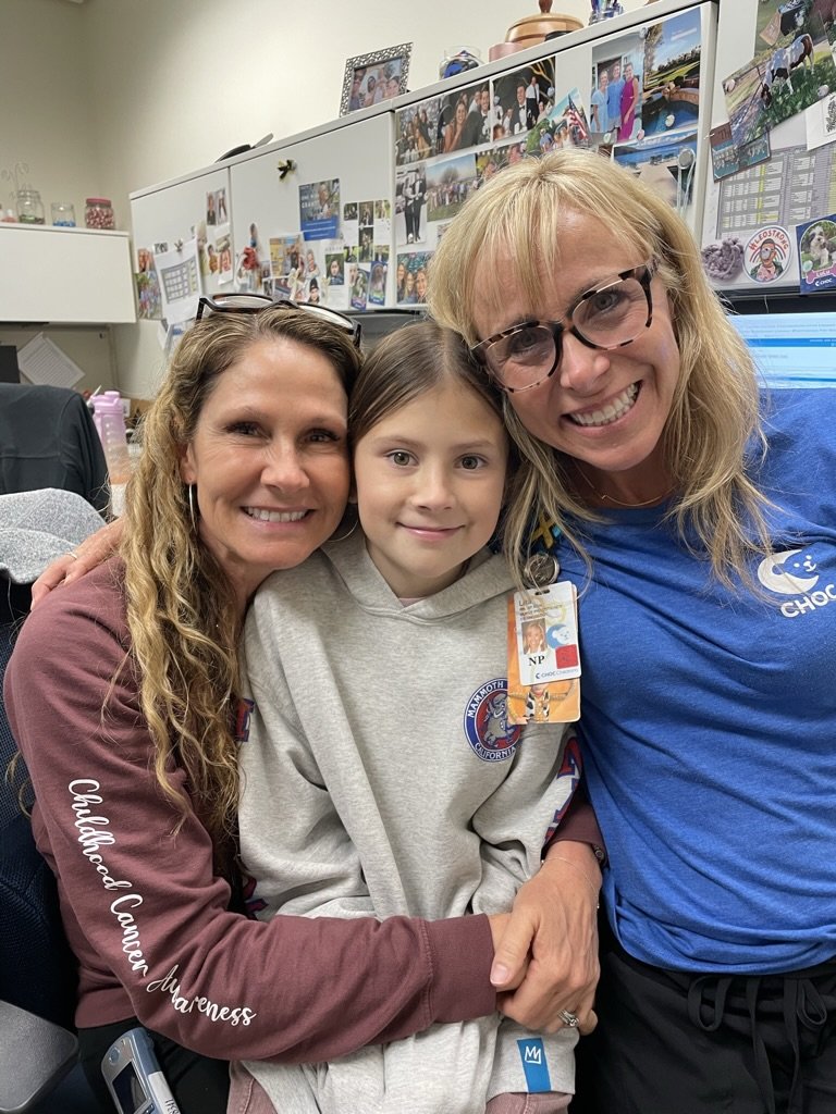 Three people smiling and hugging in an office environment, with photographs displayed on the wall behind them. One person wears a hoodie, another a maroon shirt with 'Childhood Cancer Awareness,' and the third a blue shirt.