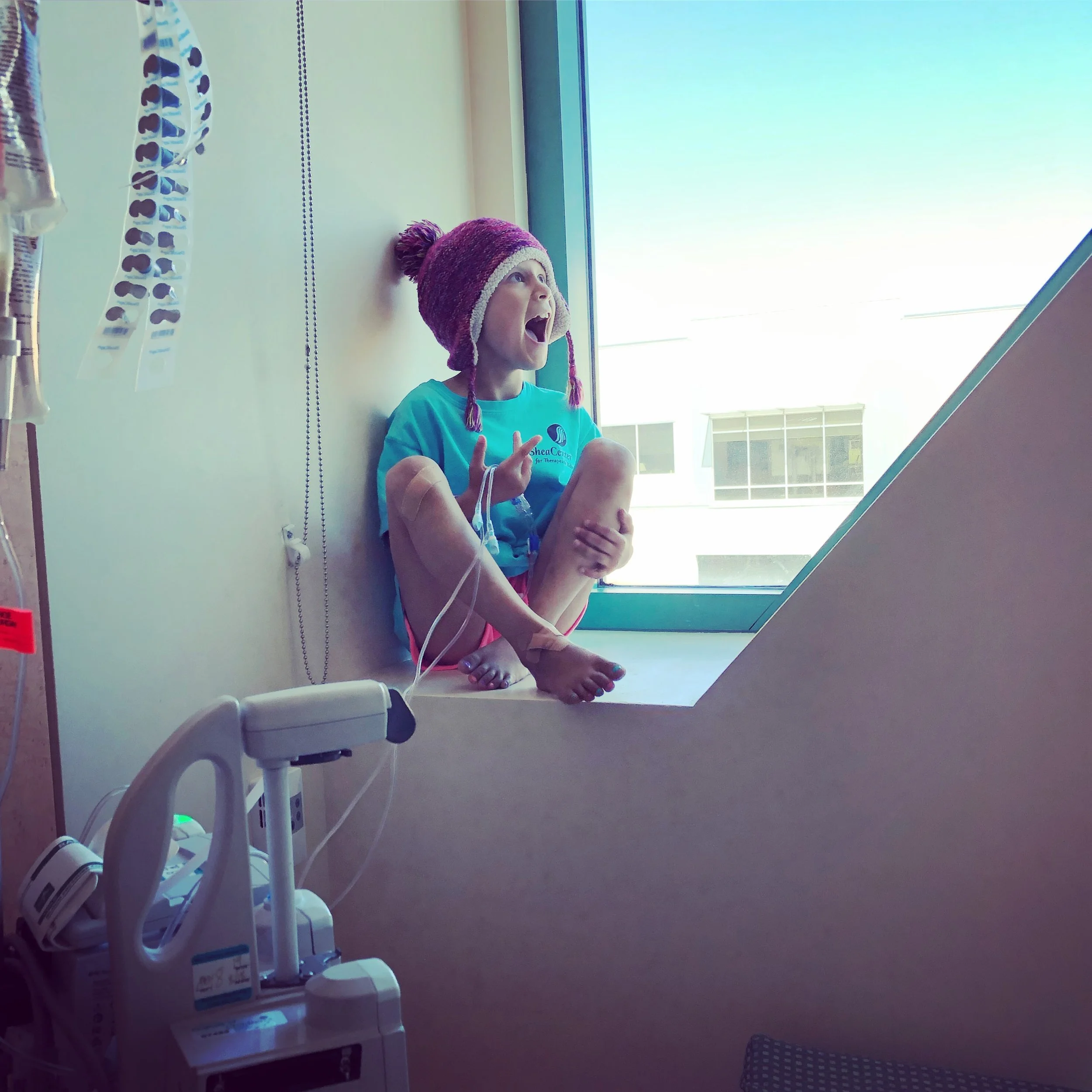 Child sitting by a hospital window wearing a colorful knit hat, connected to medical IV equipment.
