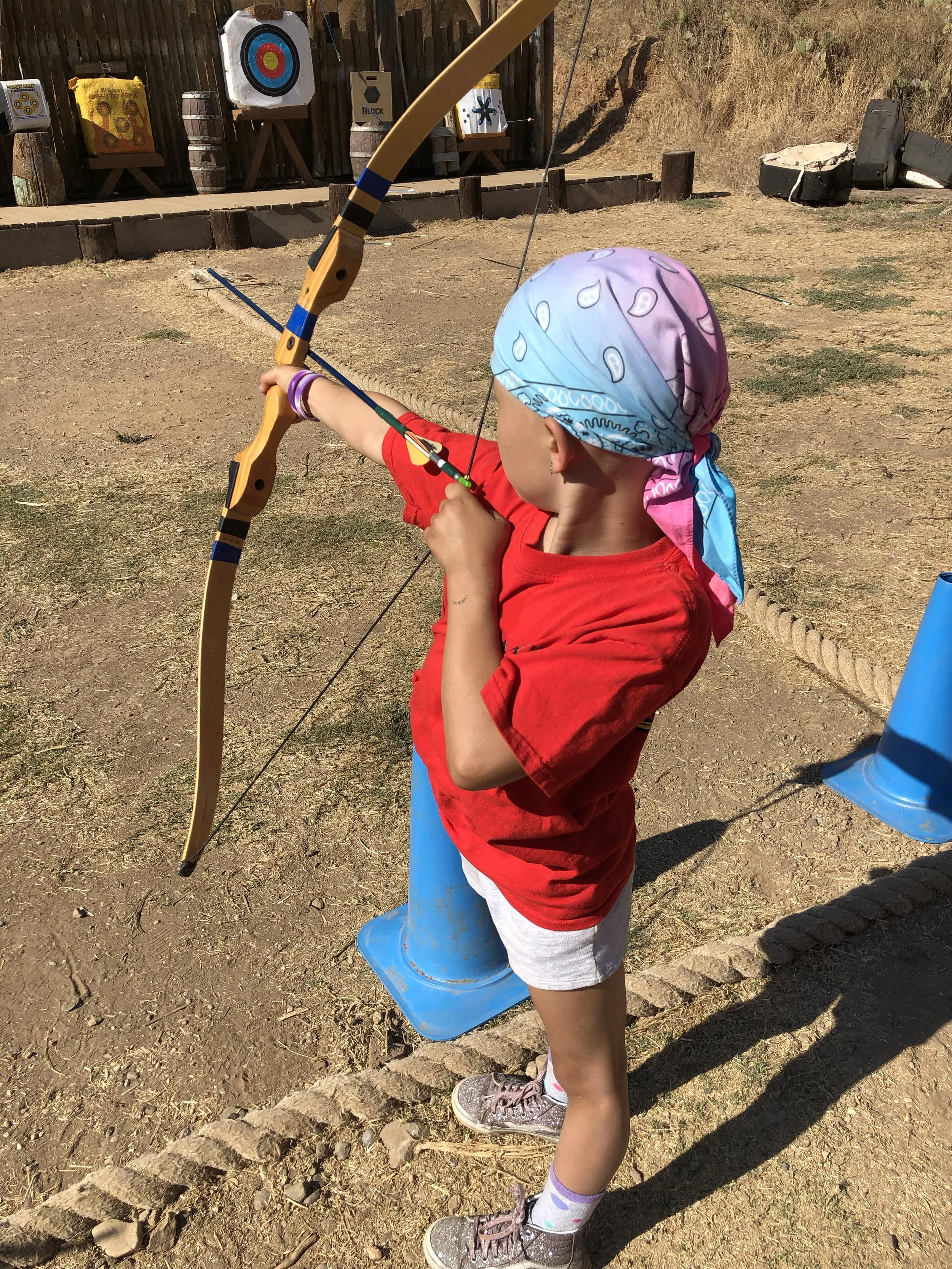 Child practicing archery at a target range, wearing a red shirt and headscarf.