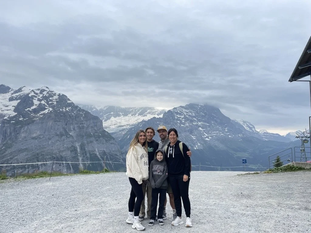 A family of five posing outdoors with snow-capped mountains in the background, under a cloudy sky.