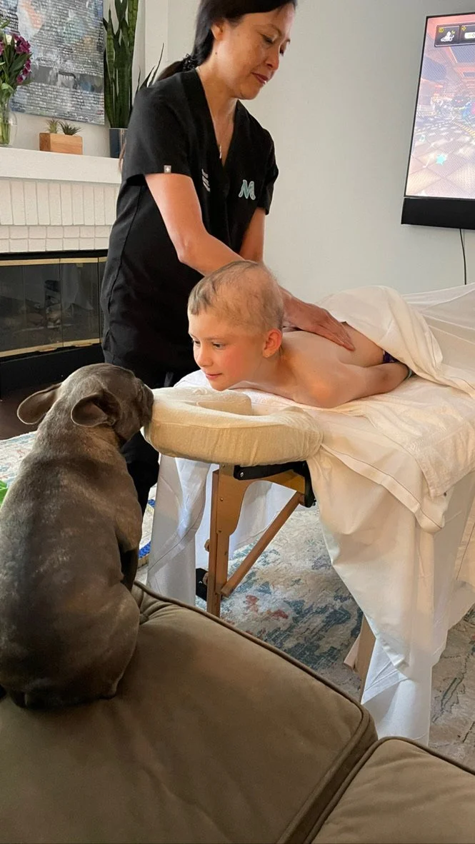 A child lying face down on a massage table receiving a massage from a massage therapist. A French Bulldog is sitting nearby, looking at the child. There's a fireplace in the background with a television to the right."
