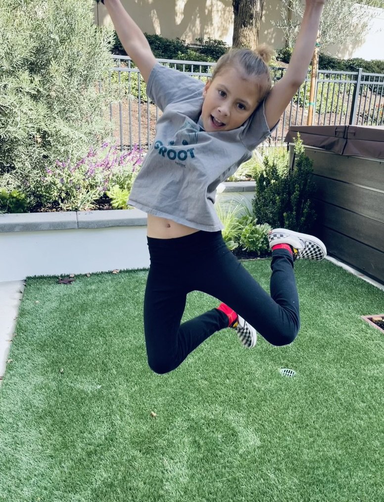 A child jumping happily on a patch of grass, wearing a gray shirt and black pants in a backyard setting with plants and a fence in the background.