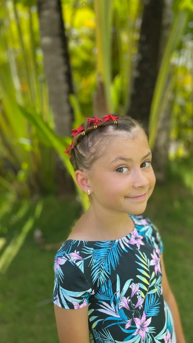 Girl with braided hair, wearing a floral dress, standing in a tropical garden.