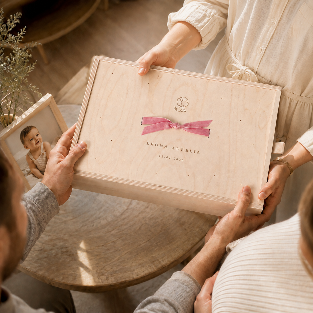 Ein Mann und eine Frau halten ein Holz-Bundles mit einer Ceremonie-Box für eine neugeborene Säugling, die den Namen "Leona Aurelia" und das Geburtsdatum 13.01.2026 trägt, ein Babyfoto liegt auf dem Tisch.