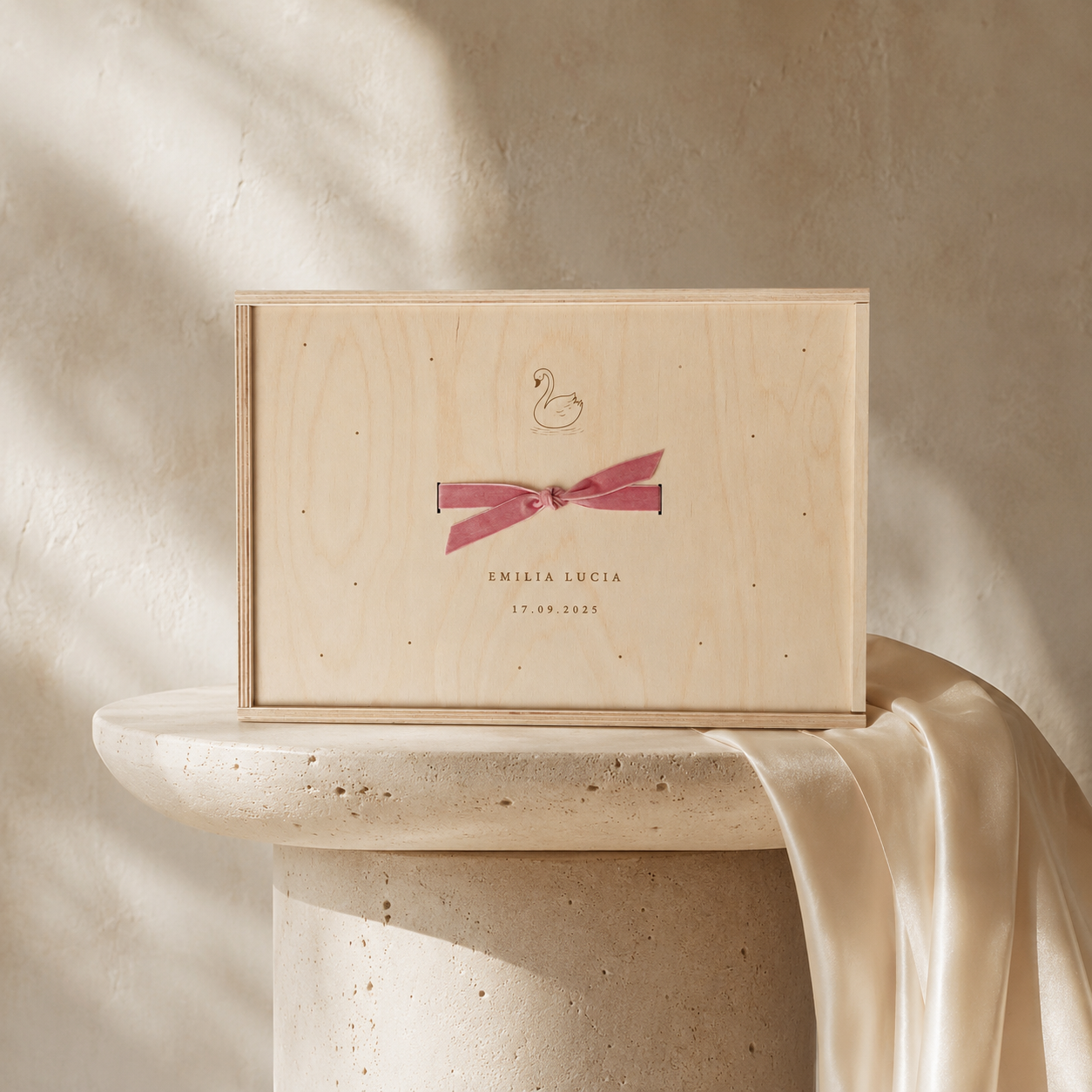 Eine Holzbox mit einer schwänenförmigen Illustration und einem rosa Band, das den Namen Emilia Lucia und das Datum 17.09.2025 trägt, steht auf einem runden, hellen Steinsockel. Der Hintergrund ist eine beige Wand mit Schatten.