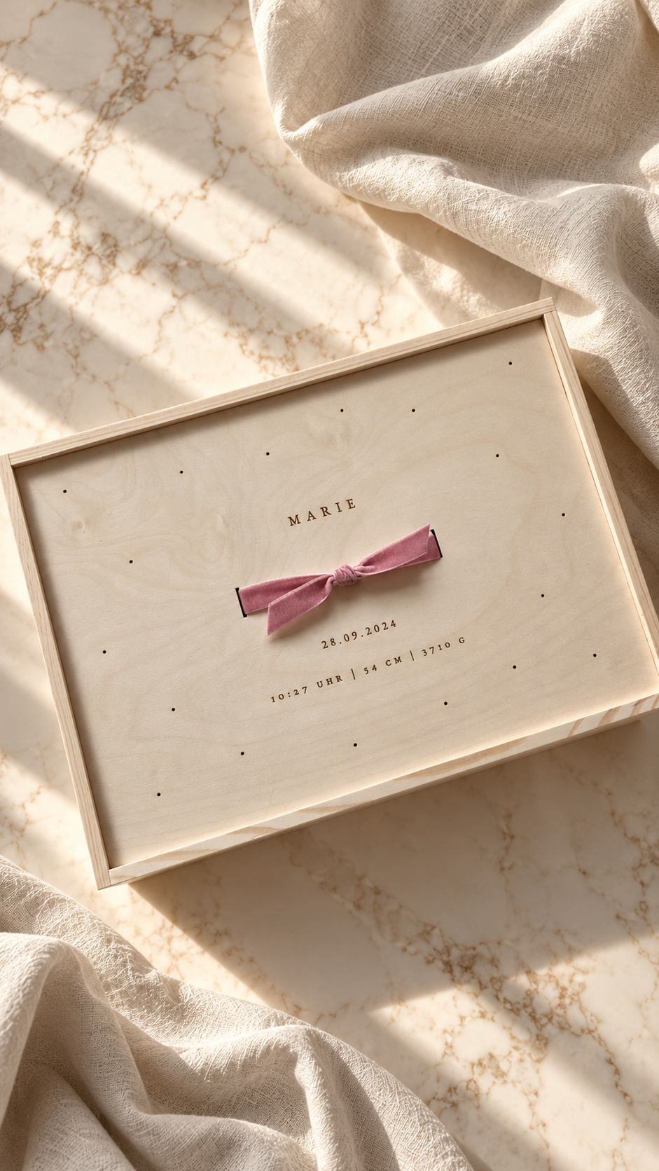 Eine Holzkiste mit einer rosa Schleife und Text, der Geburtsinformationen enthält, darunter Name, Datum, Uhrzeit, Größe und Gewicht auf einer Marmoroberfläche mit lichtdurchfluteter Kulisse.