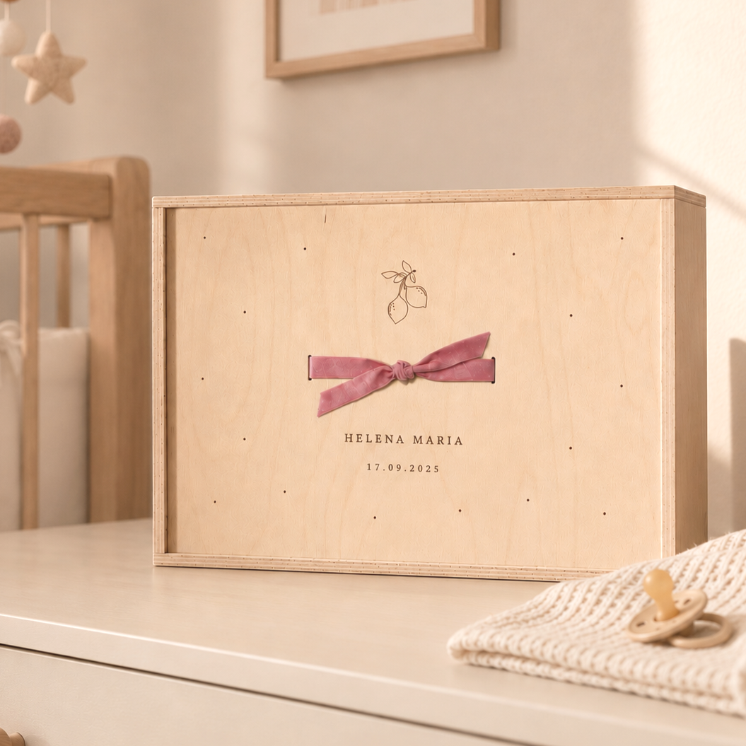 Ein Holzbox mit einer rosa Schleife und dem Namen Helena Maria sowie einem Datum, vermutlich eine Baby- oder Hochzeitsbox, auf einem weißen Möbelstück neben einem Schnuller und einem Stirnband.