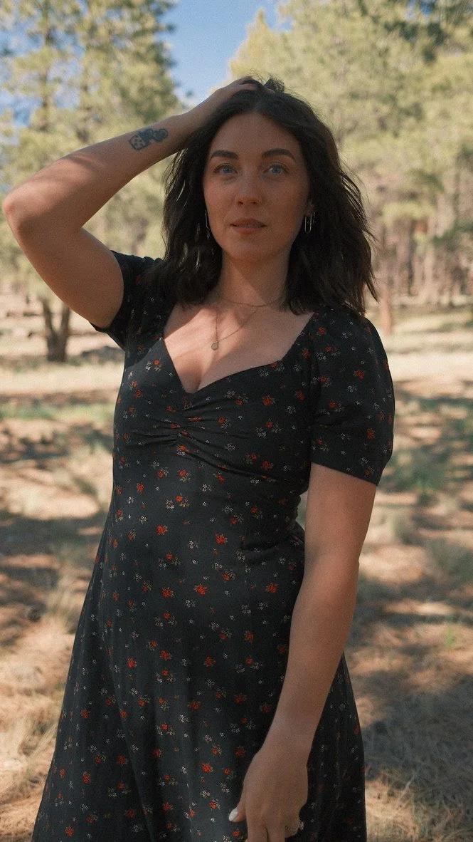 A woman standing outdoors with trees in the background, wearing a black dress with small red and white floral patterns, and holding her dark hair back with one hand.