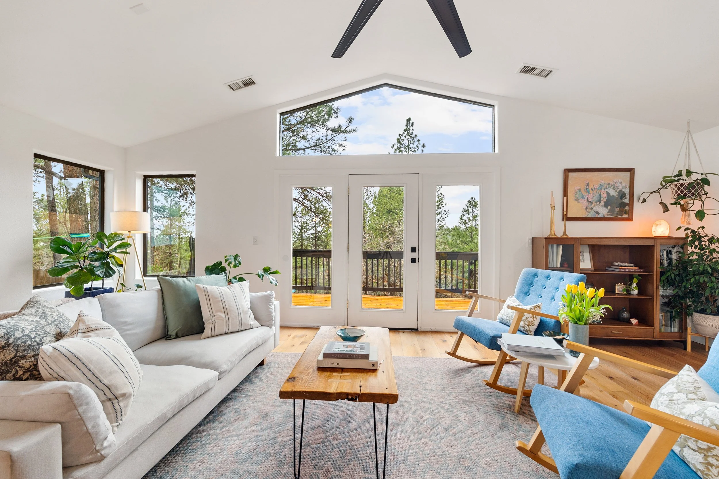 Living room with large windows showing trees outside, white walls, beige sofa with pillows, wooden coffee table with books, blue armchairs, plants, artwork, and a view of a balcony outside.