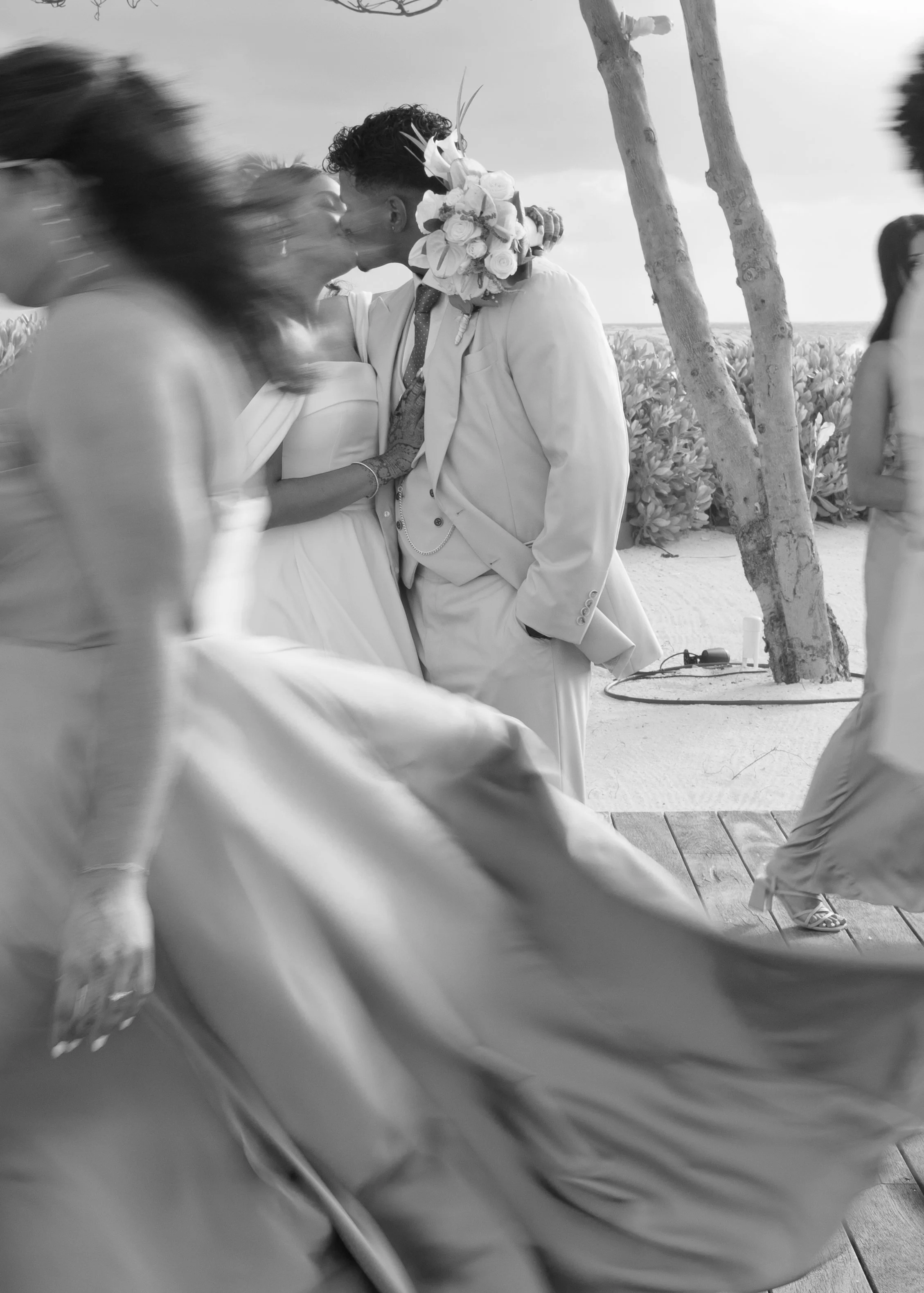 A black and white photo of a couple kissing outdoors during a sunny day, with people dancing and trees in the background.