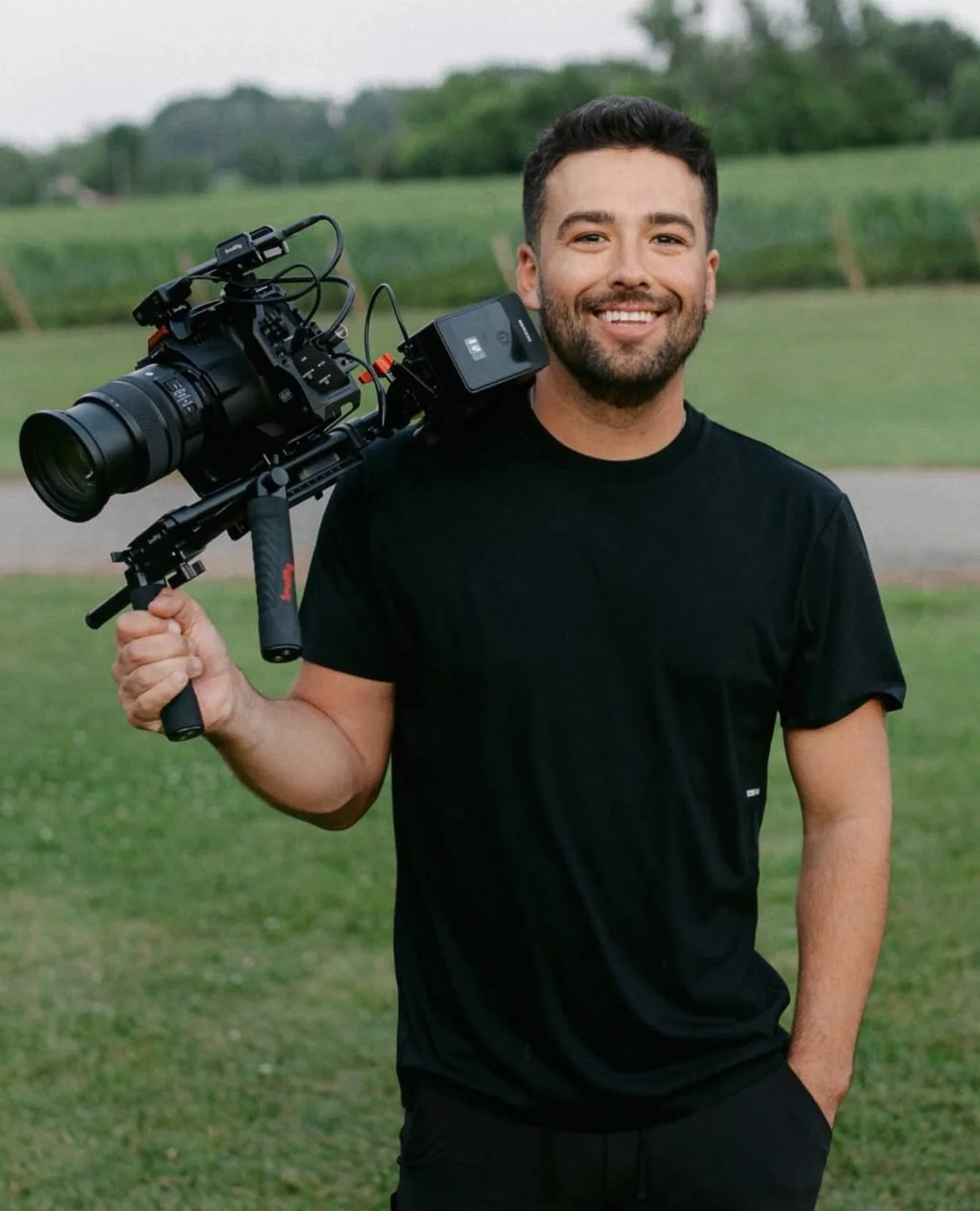 A smiling man with dark hair and a beard holding a professional video camera on his shoulder outdoors in a green field with trees in the background.