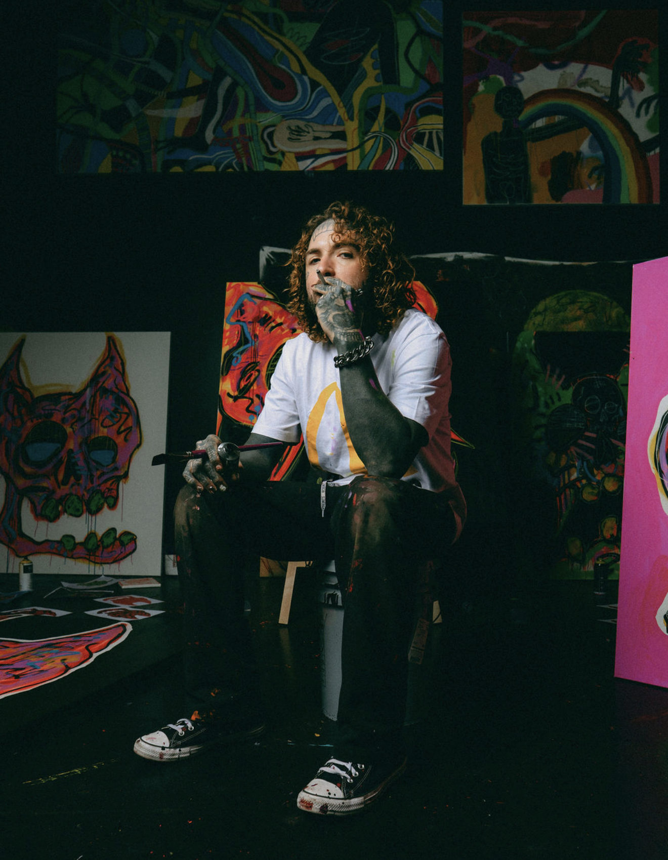 Artist Caskey seated in his studio, surrounded by colorful original paintings of skulls, figures and abstract forms.