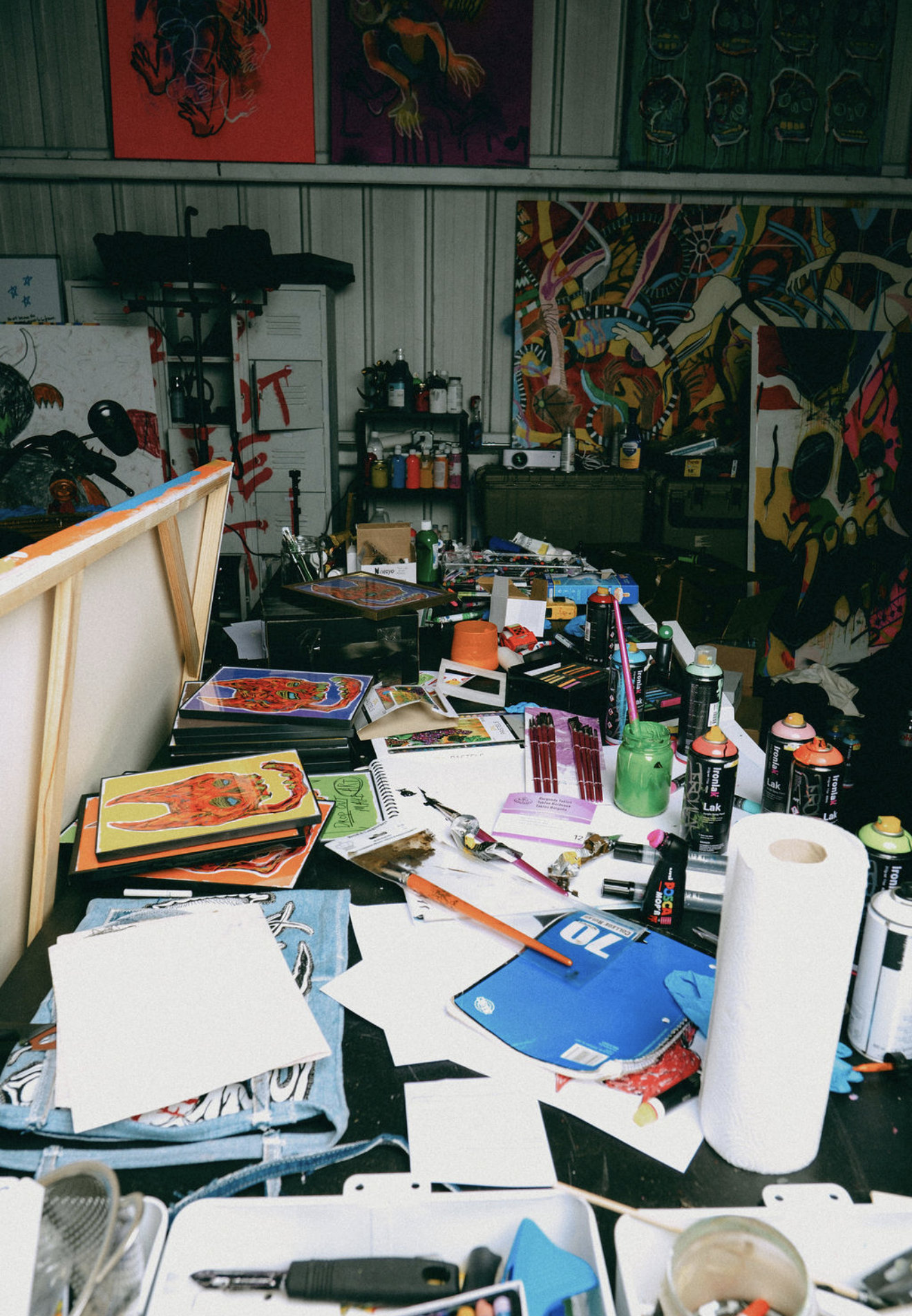 An art studio workspace cluttered with supplies, paintings, and art projects, including spray cans, paints, sketchbooks, and artwork in progress on the walls.