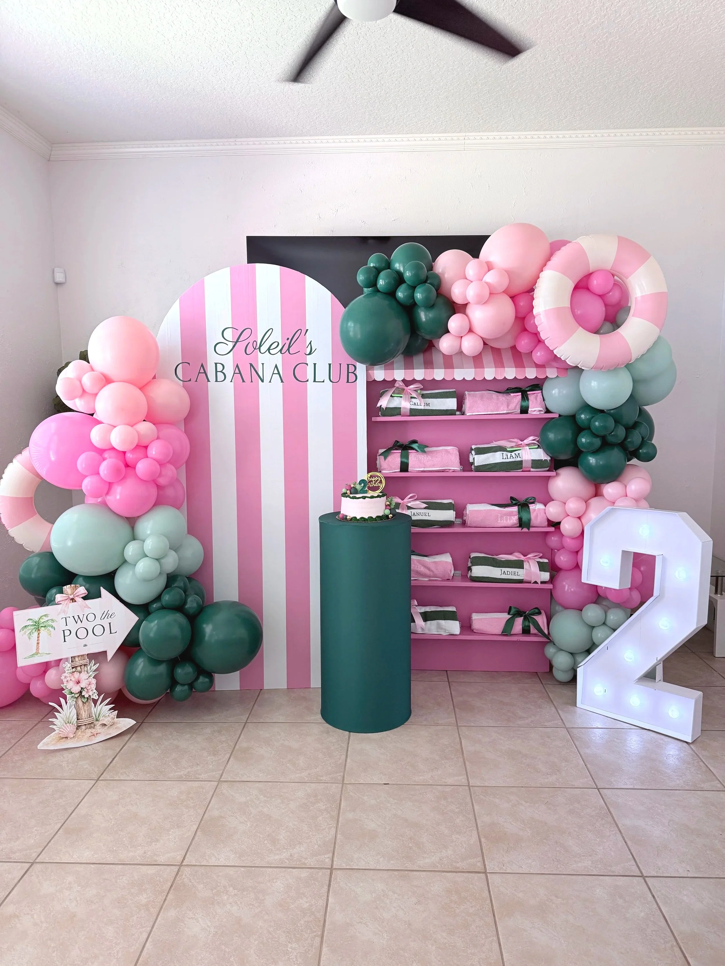 Pink and green balloon installation with a striped Joleil’s Cabana Club backdrop, cake and illuminated number two.