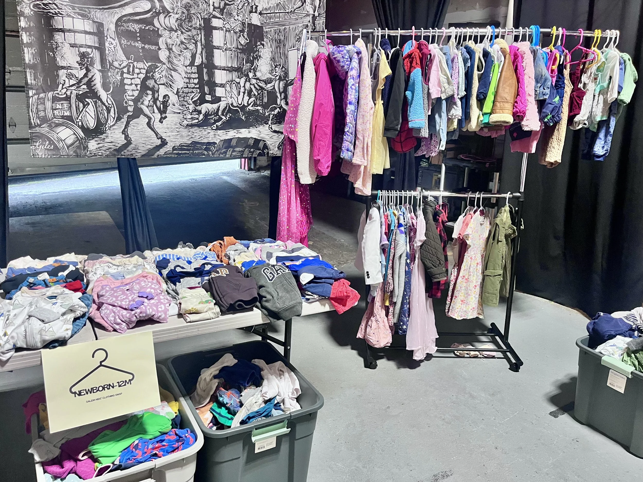 Clothing rack and containers filled with children's clothes at a sale or swap event. The rack has various kids' jackets, dresses, and outfits, with a sign indicating sizes and a lower box of folded clothes.