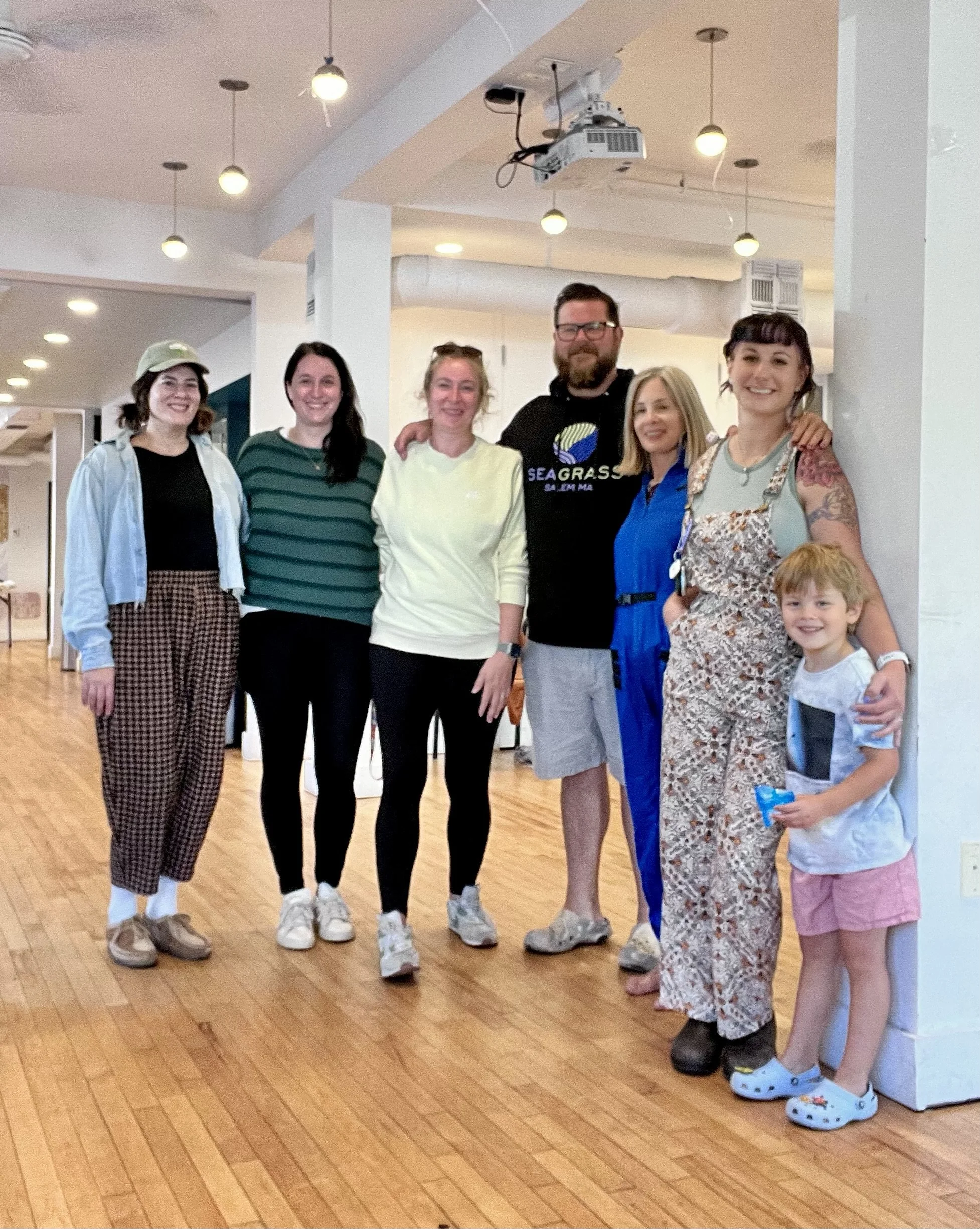 Group of six people, five women and one man, posing for a photo in a bright room. They are standing on a wooden floor, smiling at the camera. A young boy is partly visible in the lower right corner. The background includes white walls, ceiling lights, and some equipment hanging from the ceiling.