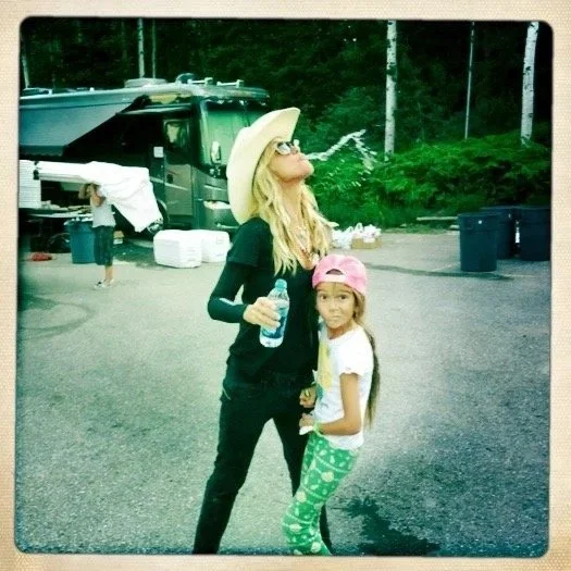 A woman wearing a large sun hat and sunglasses drinks water while standing next to a young girl with long hair wearing a pink cap and green patterned leggings. They are outdoors with a black vehicle and others in the background.
