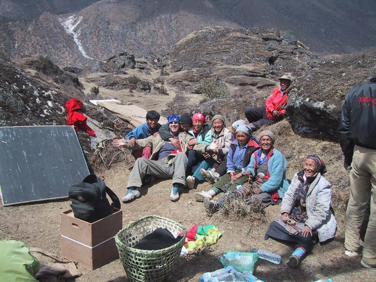 Group of hikers sitting and smiling in a mountainous, rocky landscape, surrounded by outdoor gear and supplies.