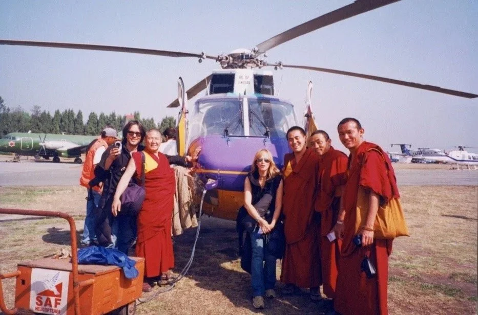 Group of seven people, including three monks in red robes and a blonde woman, standing in front of a helicopter on an airfield, with airplanes in the background.