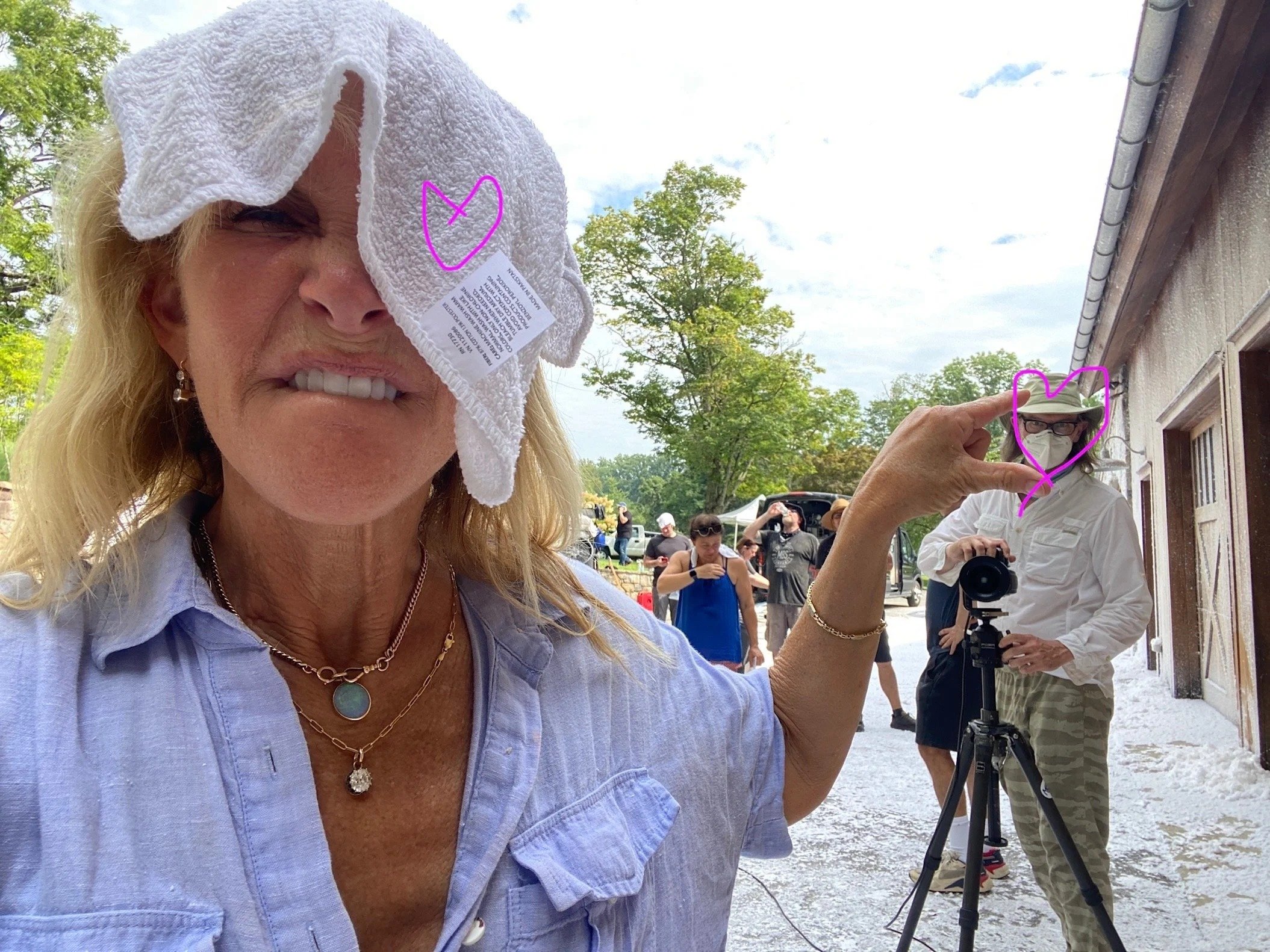 A woman with blonde hair and light skin holds a white towel over her forehead, partially covering her face. She is wearing a light blue shirt and layered necklaces, one with a round turquoise pendant. In the background, a person with glasses, a mask,