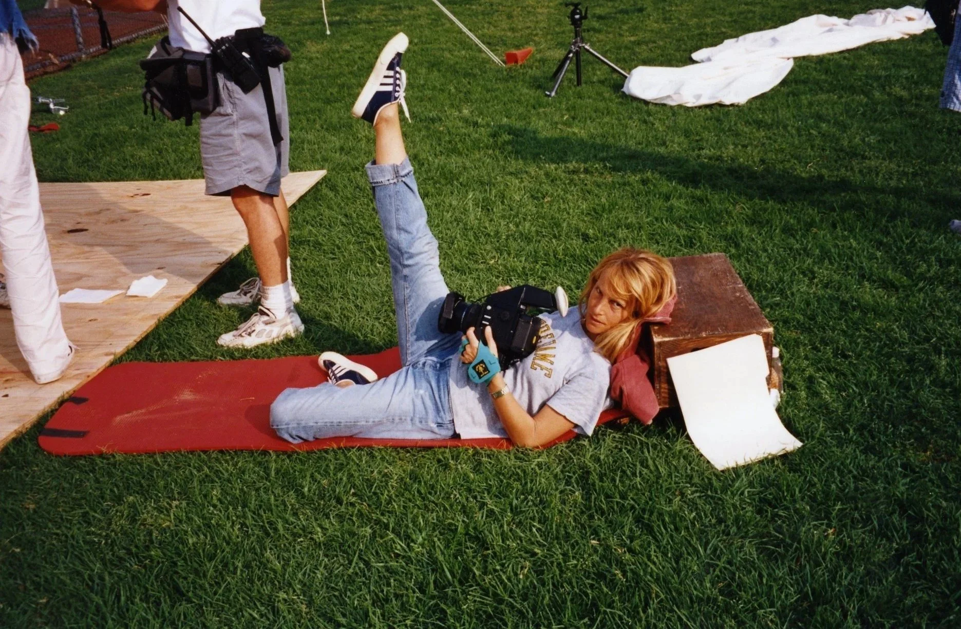 A woman lying on her side on a red mat in a grassy area, holding a camera, with her head propped on a pillow, and her leg raised. She is wearing a gray T-shirt with yellow text, jeans, and sneakers. Nearby, there are people standing and setting up eq